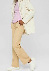 Beige Hosen mit schlankem Schnitt und leichtem Schlag; kombiniert mit einem lavendelfarbenen Ripp-Top und einer hellen cremefarbenen Jacke. Weiße Sneakers runden den Look ab.