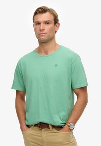 Lichtgroen katoenen t-shirt met een ronde hals en korte mouwen, voorzien van een subtiel logo op de borst, gecombineerd met kaki broek en een bruine riem.