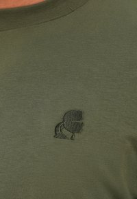 Tamnozeleni majica s kratkim rukavima s malim izvezenim crnim logom kacige na prsima.