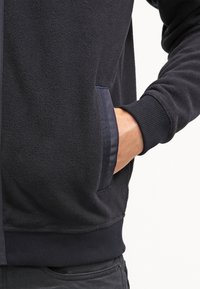 La mano di una persona nella tasca di una giacca in pile nera con polsini a coste e un dettaglio della tasca rifinito in tessuto.