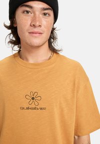 Mladá osoba s pihami, která nosí černou čepici a hořčicově žluté tričko Quiksilver s květinovým logem na bílém pozadí.
