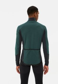 Haut de sport à manches longues en vert foncé et noir, avec une poche arrière zippée, un tissu texturé et une coupe ajustée.