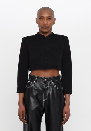 Giacca nera corta con colletto alto, zip frontale e due toppe a forma di cuore. Indossata con pantaloni neri in ecopelle a vita alta.
