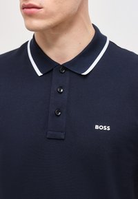 Άνδρας που φοράει μπλε ναυτικό πόλο με τρία μαύρα κουμπιά και γιακά με λευκές ρίγες, με την επωνυμία "BOSS" στο στήθος.