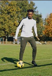 Atleet in een wit langemouwen top met zwarte accenten en donkergroene taps toelopende broek, staat op het gras en controleert een gele voetbal.