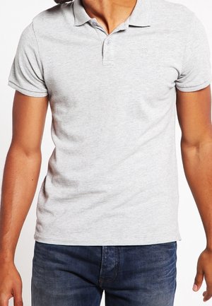 Mand iført lysegrå poloshirt med korte ærmer og mørkeblå jeans, stående mod en hvid baggrund.
