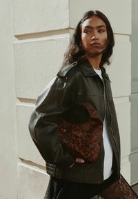 Czarna skórzana kurtka z zapięciem na zamek, żebrowymi mankietami i akcentami na ramionach, w połączeniu z torebką w panterkę. Modelka na tle jasnej ściany.