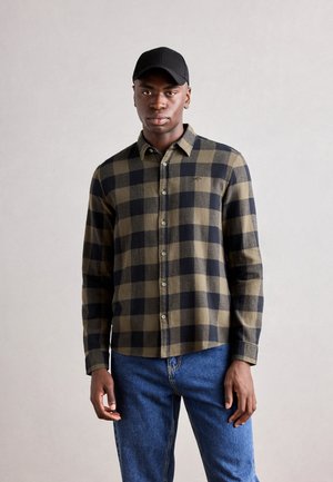 Green and black checkered flannel shirt with buttons, collared design, and long sleeves, paired with blue jeans and a black cap.