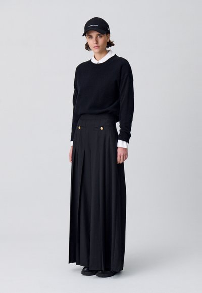 Maglione nero dal fit rilassato, camicia bianca con collo sotto, gonna plissettata nera a vita alta e cappello nero con testo bianco.