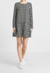 Schwarzes Langarmkleid mit weißem Blumenmuster, ausgestelltem Rock, rundem Ausschnitt und weichem Stoff. Kombiniert mit weißen Sneakers.