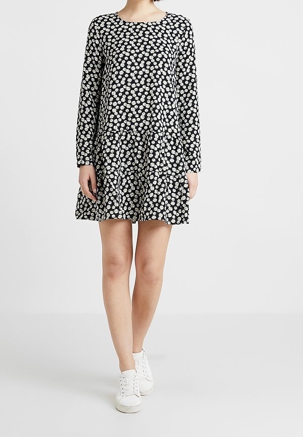 Schwarzes Langarmkleid mit weißem Blumenmuster, ausgestelltem Rock, rundem Ausschnitt und weichem Stoff. Kombiniert mit weißen Sneakers.