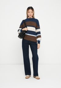 Pull rayé en mélange de laine marron, bleu et crème. Jean large en denim foncé. Le mannequin tient un sac noir et se tient contre un fond blanc.