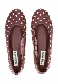 Zapatos planos burdeos con una textura suave, que presentan tachuelas plateadas y un diseño de malla con lunares. El interior tiene una plantilla acolchada y una etiqueta con el logotipo.