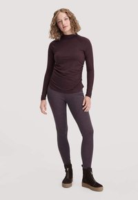 Een vrouw staat rechtop, gekleed in een donkerpaarse lange mouwen top, bijpassende legging en zwarte enkellaarzen met rits, tegen een effen achtergrond.