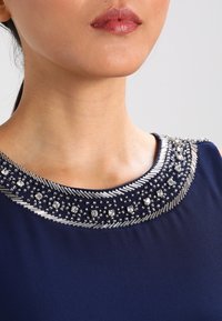 Granatowa sukienka z dekoltem wysadzanym koralikami, w tym srebrnymi koralikami i metalicznymi akcentami, podkreślająca teksturowane wykończenie i elegancki fason.