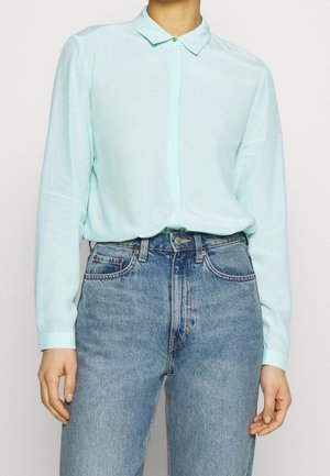 Personne portant une chemise bleu clair à manches longues boutonnée rentrée dans un jean en denim bleu taille haute avec un bouton visible et des poches.