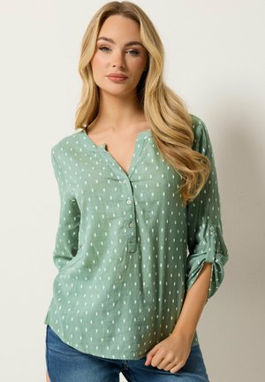 Kvinde med langt blondt hår iført en grøn skjorte med hvide rektangulære mønstre og opkrullede ærmer, stående mod en lys baggrund.