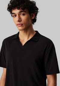 Polo nero con scollo a V, maniche corte e tessuto strutturato. Piccolo logo appare nell'area del petto sinistro.
