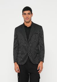 Mees, kes kannab musta bleiserit geomeetrilise mustriga, musta särgi ja mustade pükstega, seisab valge tausta ees.