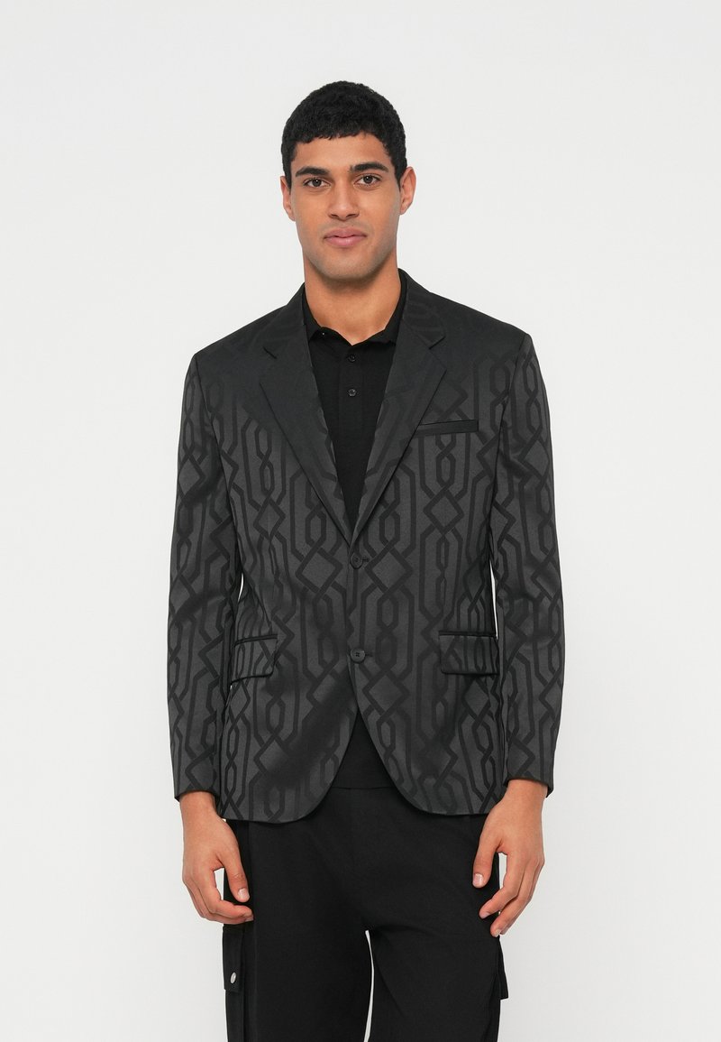 Mees, kes kannab musta bleiserit geomeetrilise mustriga, musta särgi ja mustade pükstega, seisab valge tausta ees.