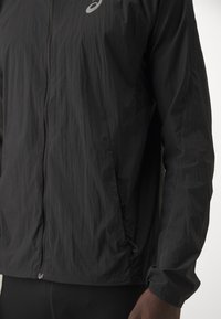 Veste légère noire zippée portée par une personne, avec des manches longues et un petit logo blanc sur la poitrine.