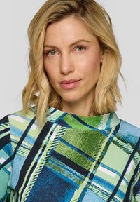 Blonde Frau mit schulterlangem Haar, die ein mehrfarbiges, grün-blau gemustertes Oberteil trägt, schaut nach vorne mit einem neutralen Ausdruck.