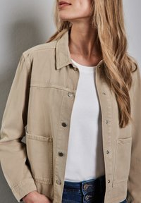 Veste beige en tissu mélange coton avec col, boutons-pression et deux poches avant, portée sur une chemise blanche et un jean bleu.