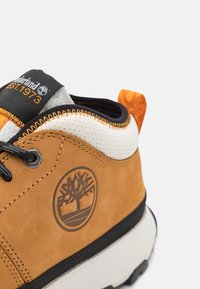 Zapatillas de ante marrón con cordones negros, cuello de malla blanca y detalles en naranja. Presenta un logo de un árbol en el lateral y una suela robusta.