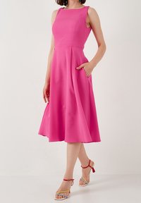 Une robe sans manches, arrivant aux genoux, de couleur rose, fabriquée en tissu lisse avec un corsage ajusté, une jupe évasée et des poches. Associée à des sandales multicolores.