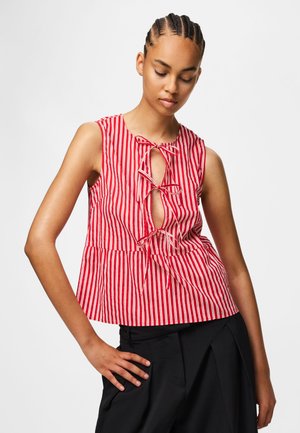 Femme aux cheveux tressés portant un haut sans manches à rayures verticales rouges et blanches avec trois nœuds papillons sur le devant, associé à un pantalon noir.