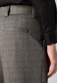 Grau karierte Hose mit subtiler Textur, einem einzelnen Gesäßtasche und Gürtelschlaufen, getragen mit einem schwarzen Langarmshirt.