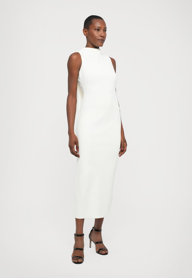 Robe midi blanche sans manches avec un design à col montant, texture lisse et fentes sur les côtés, accompagnée de chaussures à talons hauts noires à lanières.