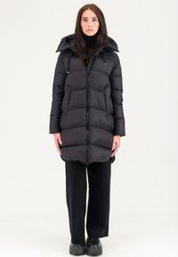 Doudoune noire avec fermeture éclair, capuche et poches avant. Présente un design matelassé et une finition mate, associée à un pantalon et des chaussures noirs.