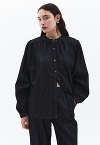 Tmavě modrá džínová košile na knoflíky se staženým límcem, dlouhými rukávy a volným střihem. Vybavena jemnou texturou a odpovídajícími kalhotami.