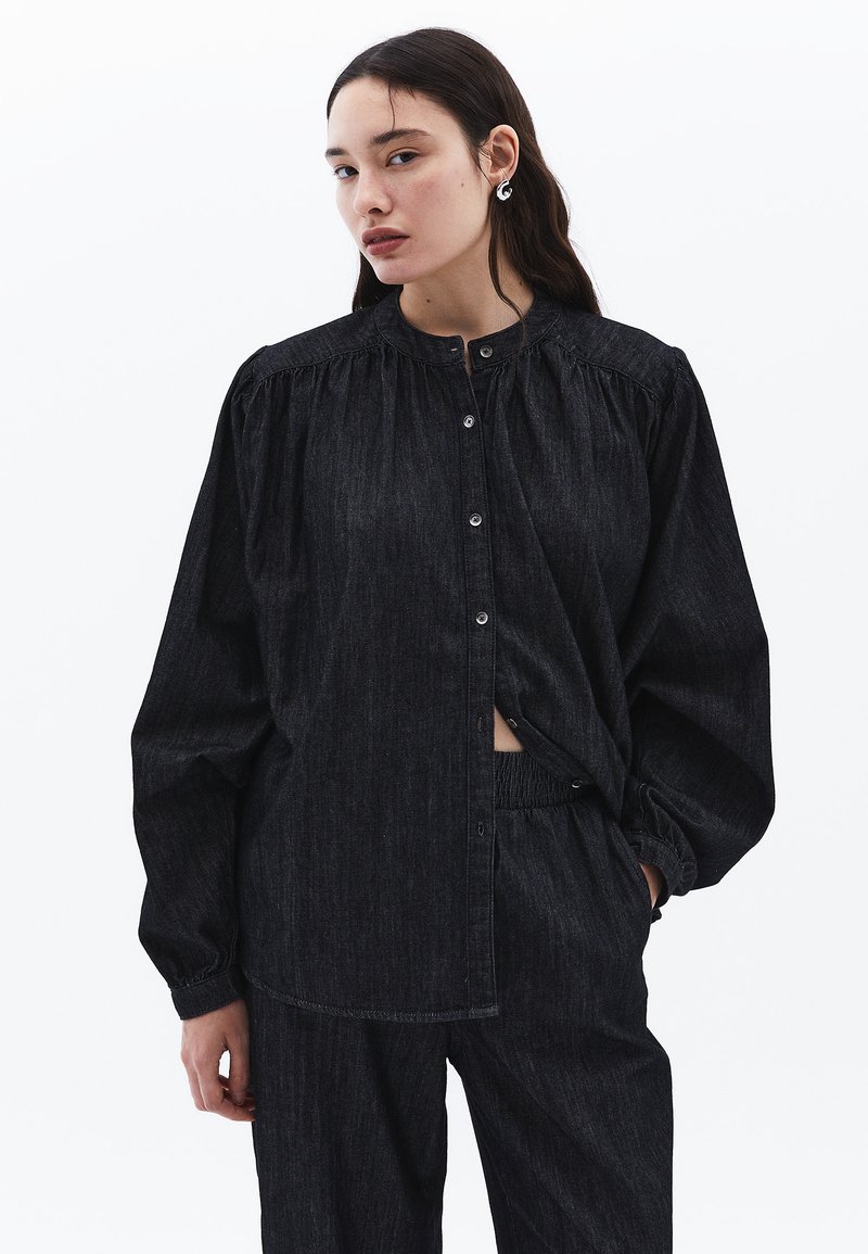 Tmavě modrá džínová košile na knoflíky se staženým límcem, dlouhými rukávy a volným střihem. Vybavena jemnou texturou a odpovídajícími kalhotami.