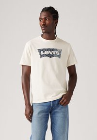 Hvit T-skjorte med rund hals, med et marineblått mønstrete Levi's-logo på fronten, kombinert med lyseblå denimjeans.