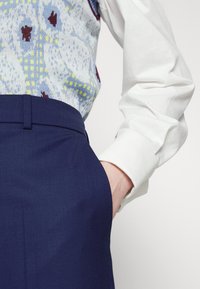 Personne portant un pantalon bleu marine et une chemise blanche à manches longues avec un gilet à motifs, main placée dans la poche du pantalon.