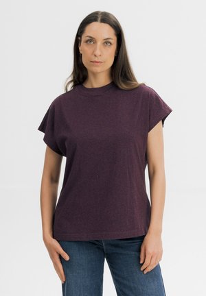 Vrouw met lang donker haar, die een paarse korte mouwen top en een blauwe spijkerbroek draagt, staand tegen een effen lichte achtergrond.