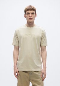 Bézs pamut póló kerek nyakkivágással, előoldalán dombornyomott "BOSS" logóval. Rövid ujjú, laza szabású, letisztult dizájn.