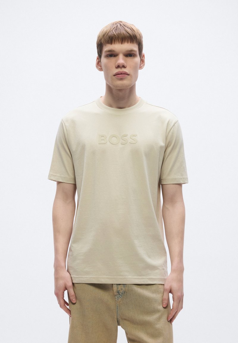 Bézs pamut póló kerek nyakkivágással, előoldalán dombornyomott "BOSS" logóval. Rövid ujjú, laza szabású, letisztult dizájn.