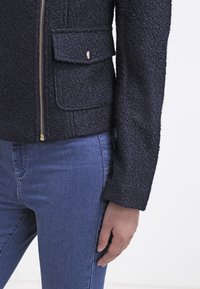 Zbliżenie na osobę w teksturowanej czarnej kurtce na zamek z klapką i niebieskich dopasowanych dżinsach, ręka luzno oparta na boku.