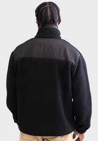 Jachetă din fleece negru cu un corp texturat, panou de umăr negru neted, guler înalt și manșete elastice. Vedere din spate cu detalii de design minime.