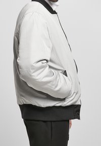 Man draagt een witte bomberjack met zwarte kraag en zoom, met de handen in de zakken en zwarte broek, zijaanzicht op heuphoogte.