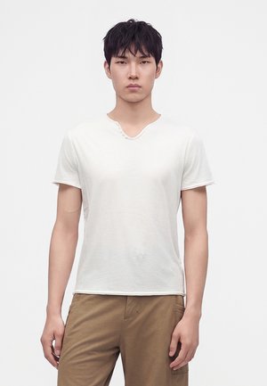 Jonge man met kort zwart haar die een effen wit shirt met korte mouwen en lichtbruine broek draagt, staand tegen een witte achtergrond.