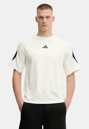 Hombre con el cabello corto, vistiendo una camiseta blanca Adidas de manga corta con detalles negros y pantalones negros, de pie con las manos en los bolsillos.