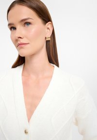 Biely pletený kardigán s hlbokým V-výstrihom, textúrovanými vzormi a zlatým gombíkovým zapínaním. Modelka doplnila svoj vzhľad zlatými kruhovými náušnicami.