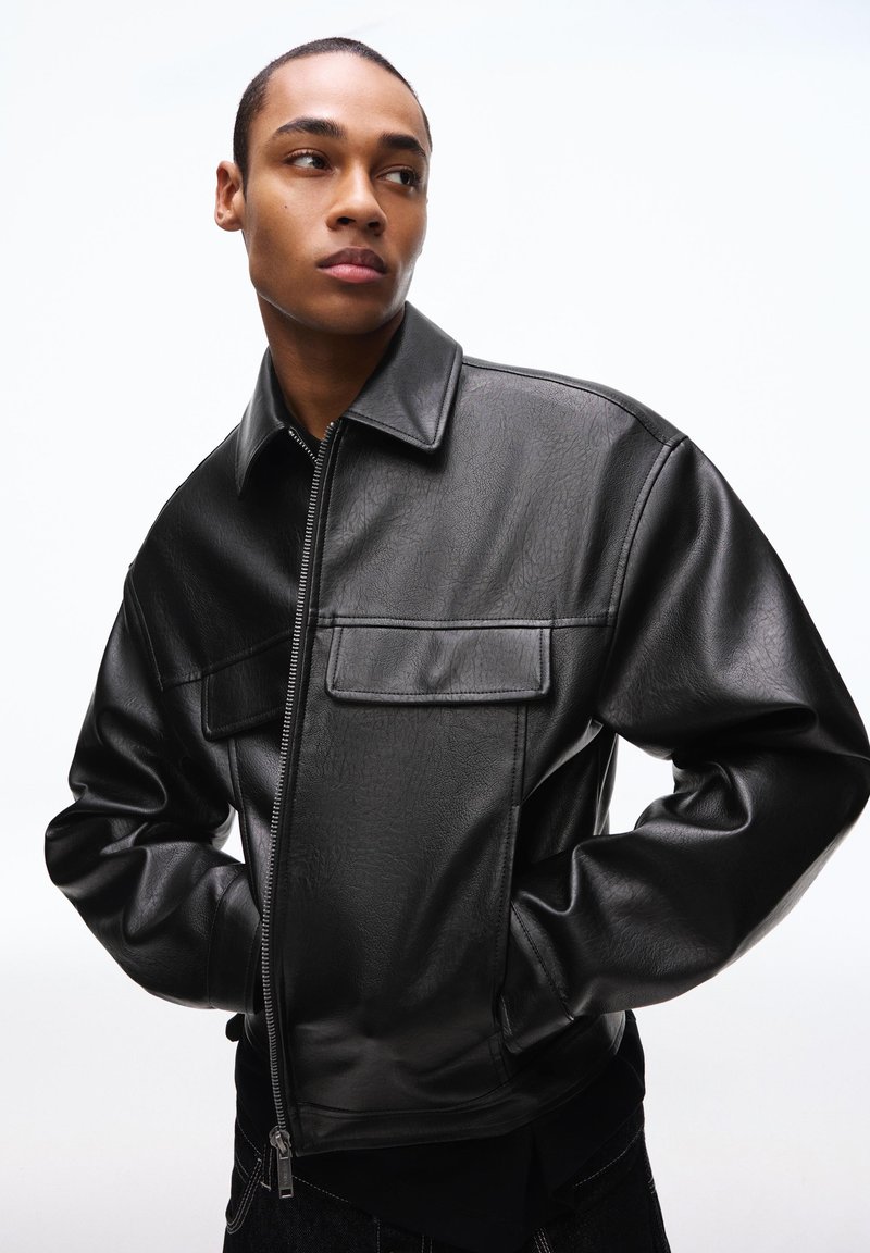 Junger Mann mit kurzen Haaren, der eine schwarze Lederjacke mit vorderen Klappentaschen trägt, mit den Händen in den Taschen steht und zur Seite schaut.