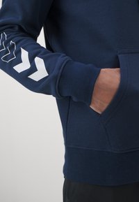 Sweat-shirt bleu marine avec une poche frontale, des poignets côtelés et trois motifs en forme de flèche blancs sur la manche gauche. Texture de tissu lisse.