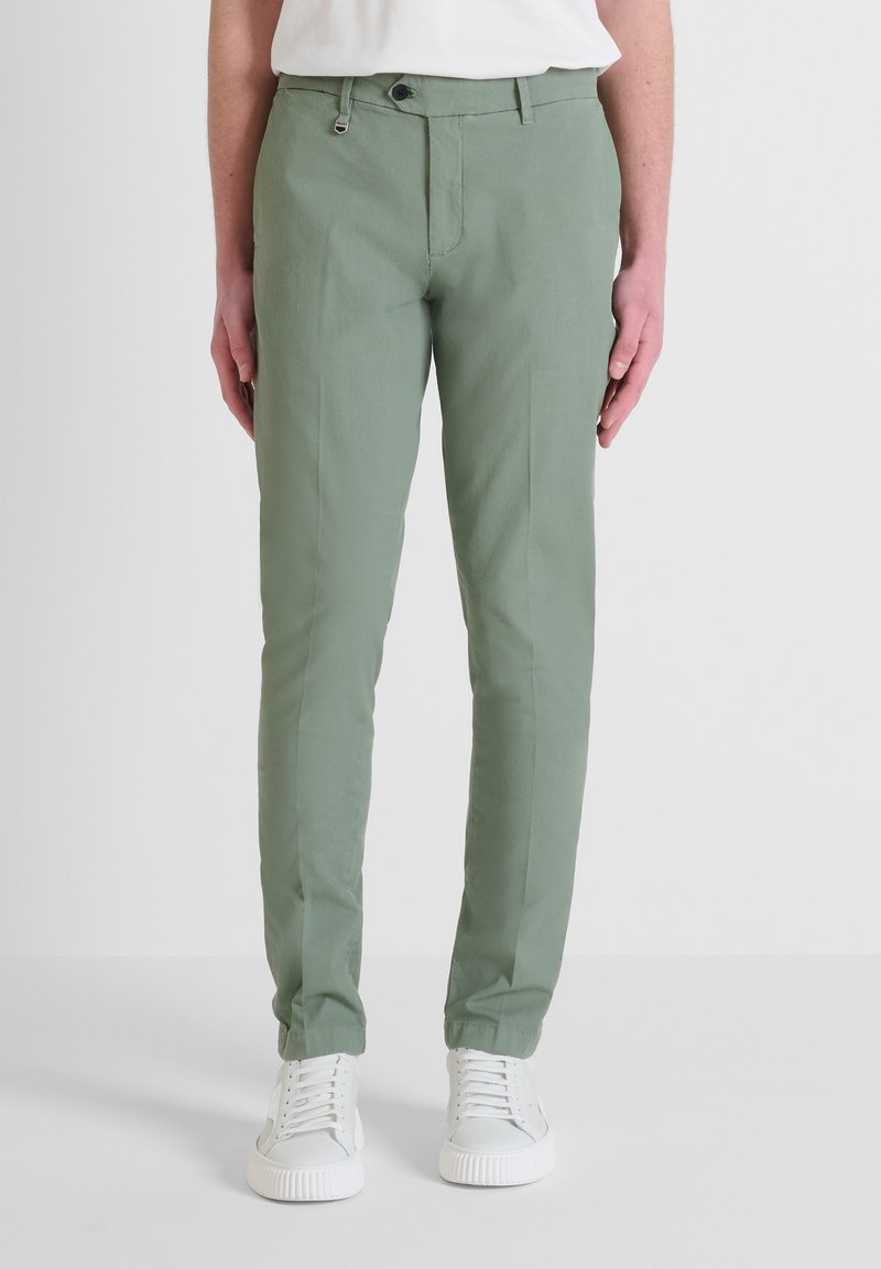 Pantaloni slim-fit di cotone verde chiaro. Presentano una parte anteriore piatta, chiusura con zip e passanti per la cintura. Indossati con sneakers bianche.