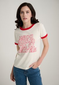 T-shirt crème à manches courtes avec col côtelé rouge, présentant un texte audacieux et inversé : « BIENVENUE DANS LE MONDE À L'ENVERS ». Porté avec un jean bleu.